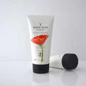 A tube of NAPA SOAP COMPANY body butter with a floral design, lying next to its cap on a white surface.