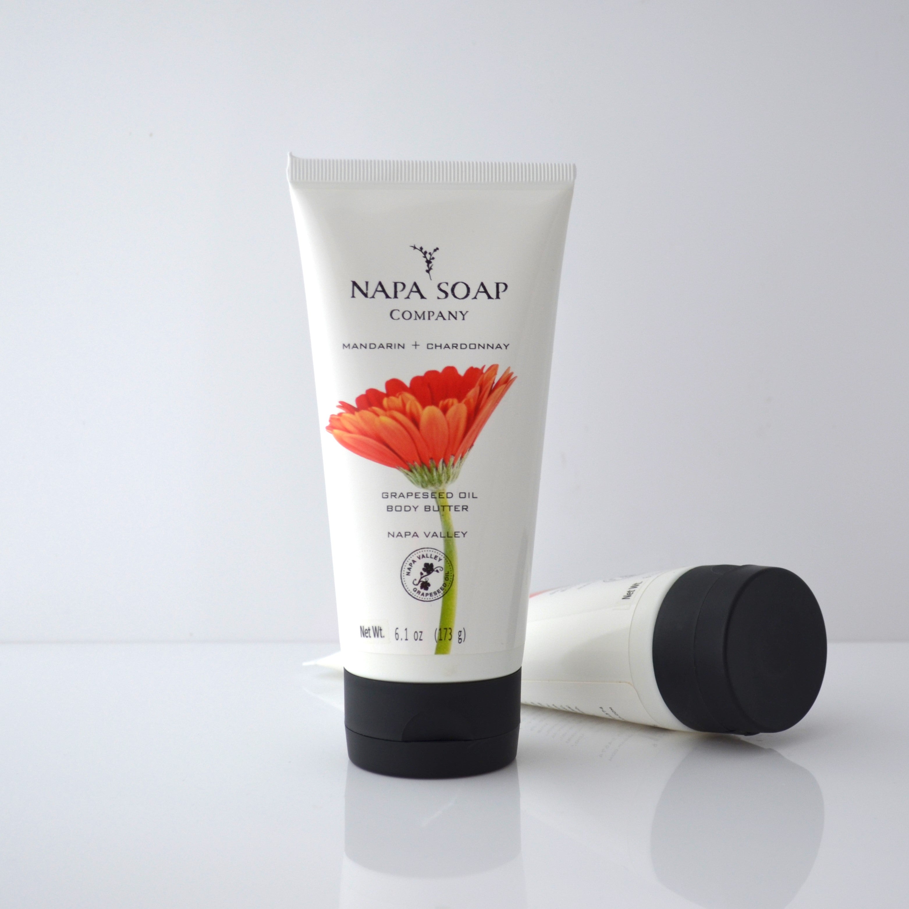 A tube of NAPA SOAP COMPANY body butter with a floral design, lying next to its cap on a white surface.
