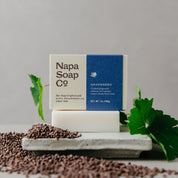 Napa Soap Co. soap bar with packaging on a gray surface with grape seeds and grape leaves.